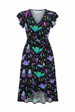 Load image into Gallery viewer, A woman modeling a short-sleeve V-neck dress with a black background and a colorful print of green, purple, and blue fairies and butterflies, holding a small woven handbag against a plain white background.